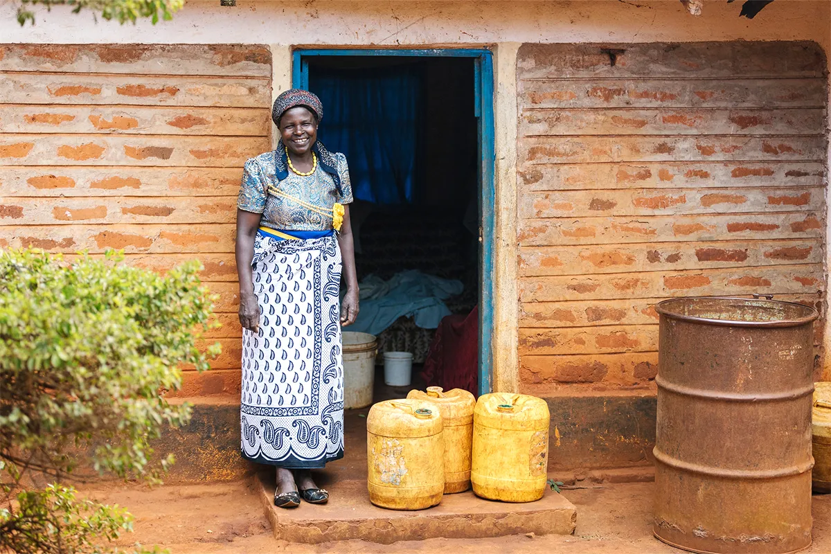 Woman standing in front of her home in Kenya with three water containers.