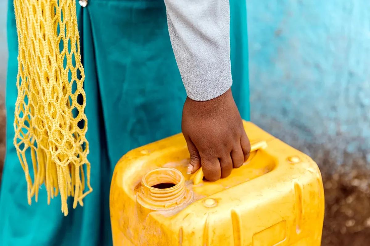 Person carrying a yellow water vessel in front of a teal wall.